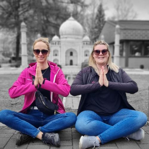 Zwei Frauen sitzen vor dem Taj Mahal. Die Personen sind in Farbe und falten die Hände, der Hintergrund  in schwarz-weiß