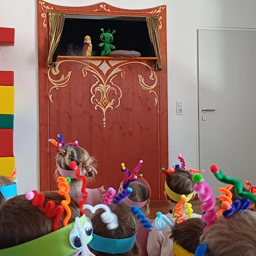 Auf der Kasperlbühne sind der Kasperl und ein Alien zu sehen. Die Kinder mit ihrer Alien- Kopfbedeckung beobachten die Handlung