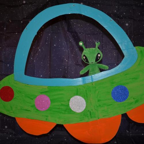 Ein Stoffalien schaut aus einem selbstgebastelten Raumschiff heraus. Den Hintergrund bildet ein Sternenhimmel