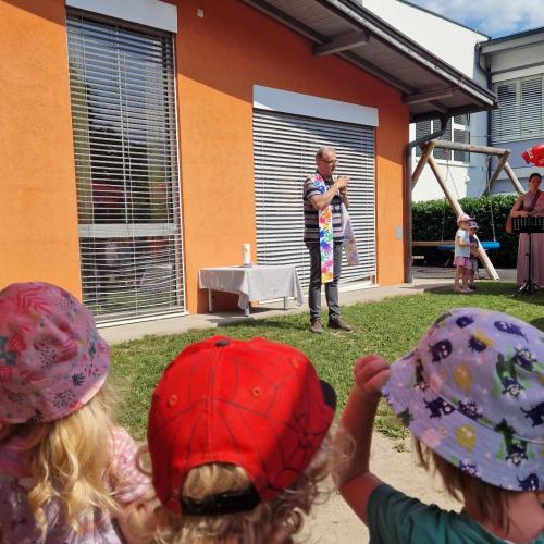Pfarrer Josef steht im Garten des Kindergartens und spricht zu den Kindern, die im Halbkreis um ihn stehen