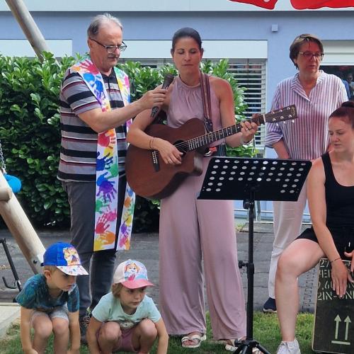 Die Pädagogin spielt stehend mit der Gitarre, Neen ihr spielt eine Päd. auf der Cajon und steht die Frau Bgm. Der Pfarrer hält das Mikrofon 