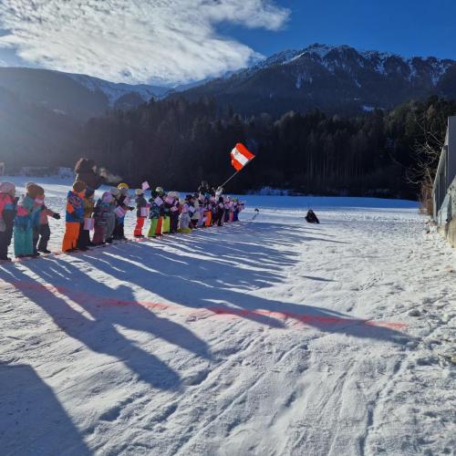 Rutschblattrennen, ein Kind rutsch den Hang herunter und wird an der Seitenlinie von den anderen mit Fahnen und Zurufen unterstützt