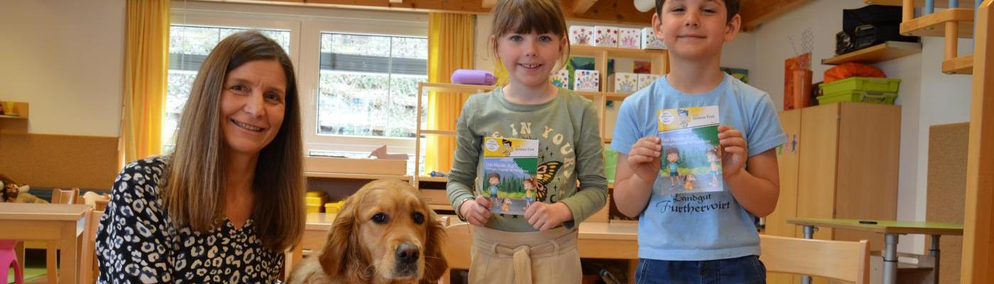 Ein Therapiehund mit seiner Begleiterin und zwei Kinder die ein Sicherheitsbüchlein in der Hand halten