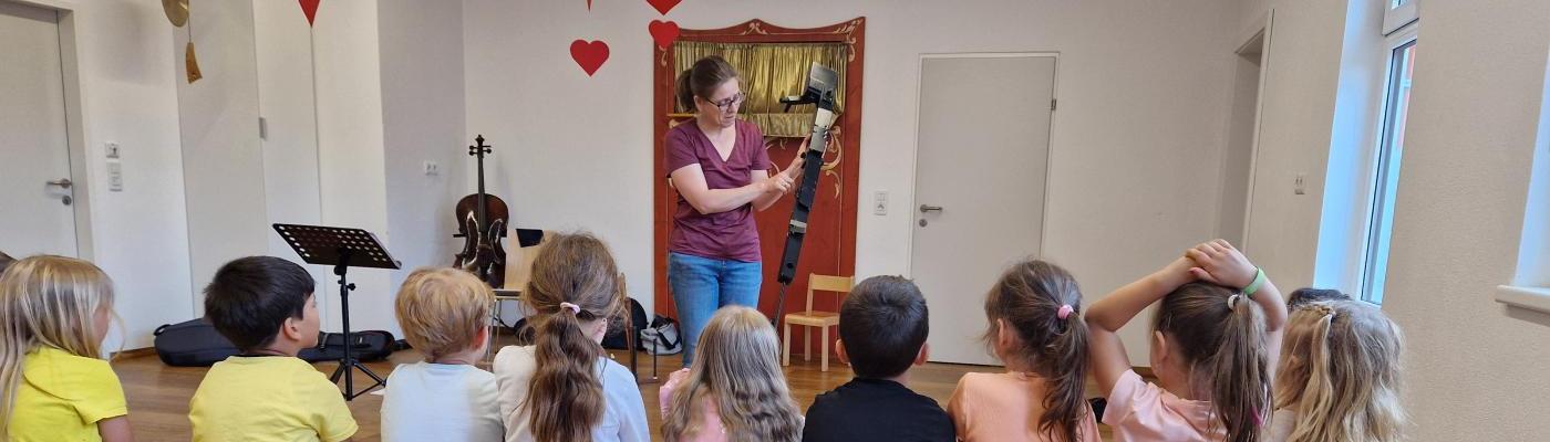 Die Kinder auf einer Langbank sitzend von hinten fotografiert. die Musiklehrerin hält eine Flöte in der Hand 