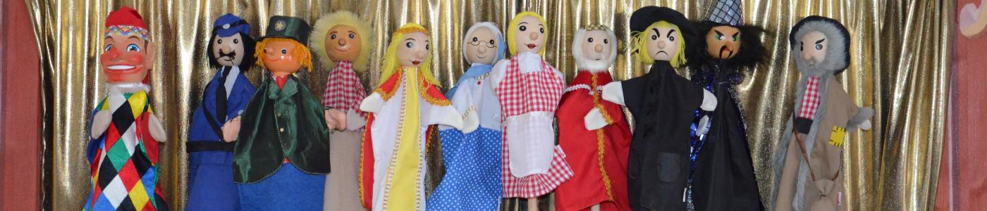 Viele Kasperlfiguren stehen nebeneinander auf einem Ständer in der Bühne des Kasperltheaters vor dem goldenen Vorhang