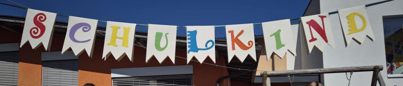 Szene Rausschmeisserfest, Alle Kinder des Kindergartens stehen und beobachten wie die vier Pädagoginnen Schulkinder rausschmeissen. Sie halten sie bei den Füßen und unter den Achseln und "werfen" sie auf die gr. Pölster die unter dem Schulkind-Banner liegen