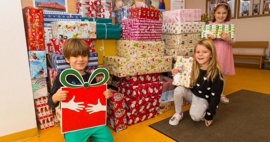 3Kinder stehen mit Geschenken vor den vielen Weihnachtpäckchen die gespendet wurden