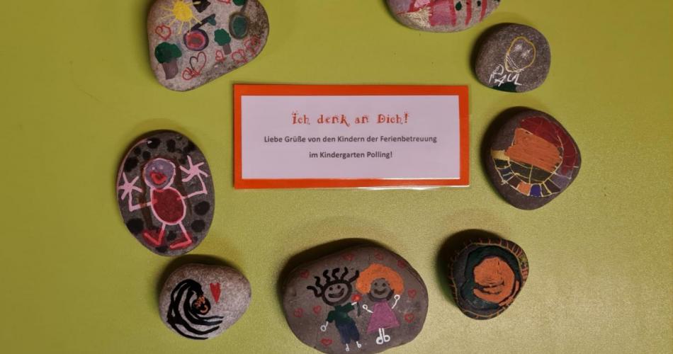 Von Kindern bemalte Steine liegen in einem Kreis um dem Text "Ich denk an dich. Liebe Grüße von den Kindern der Ferinbetreuung"