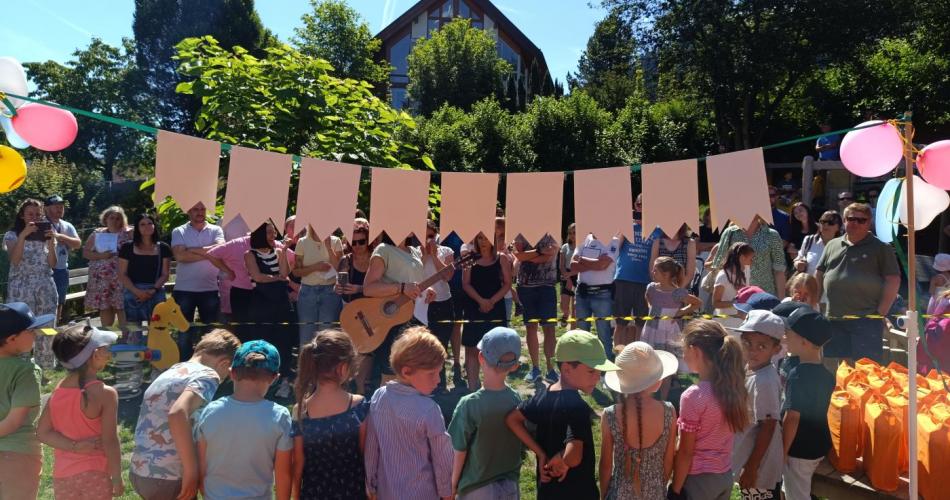 Die 18 Schulkinder stehen mit dem Rücken zum Foto unter der Schulkind Girlande. Im Hintergrund stehen Eltern die extra für das Rausschmeißerfest gekommen sind