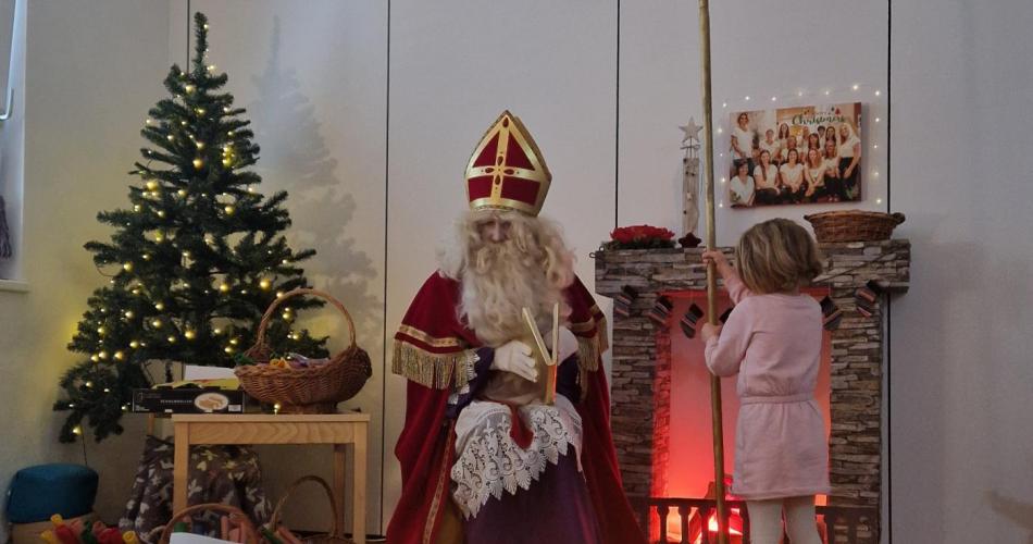Der Nikolaus sitzt vor dem beleuchtenden Tannenbaum und ein Kind hält seinen Nikolausstab