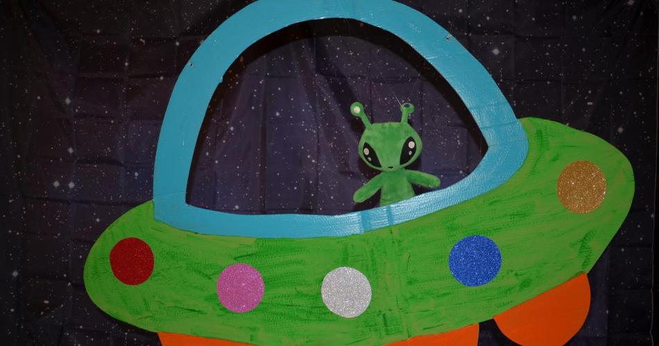 Ein Stoffalien schaut aus einem selbstgebastelten Raumschiff heraus. Den Hintergrund bildet ein Sternenhimmel