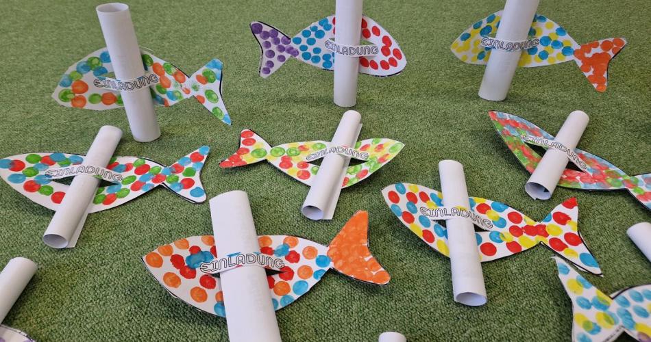 Einladungskarten von den Kindern gebastelt. Ein einfacher Fisch wurde mit bunten Fingerabdrücken verziert und in der Mitte zwei mal eingeschnitten. Dadurch ergibt sich eine Schlaufe in der die zusammengerollte Einladung zum Fest  durchgeschoben  wurde