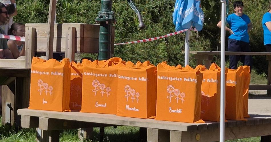 Mehrere Stofftaschen die mit dem Logo des Kindergartens und dem Namen des Schulkindes versehen sind stehen im Garten auf einem Holpodest bereit