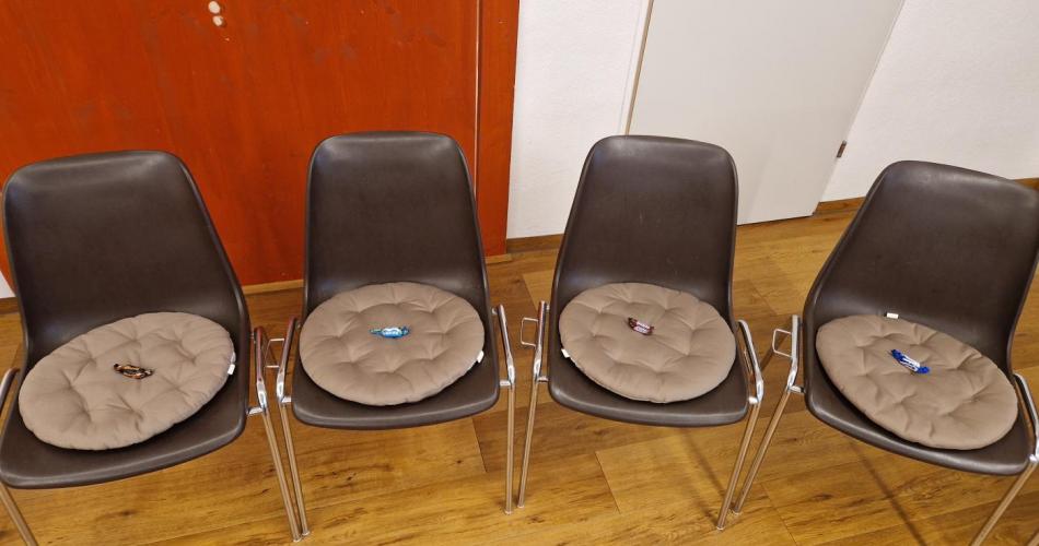Ein Ausschnitt eines Stuhlkreises. Vier Stühle mit einem Sitzkissen sind zu sehen, auf denen eine kleine Süßigkeit für den Teilnehmenden liegt