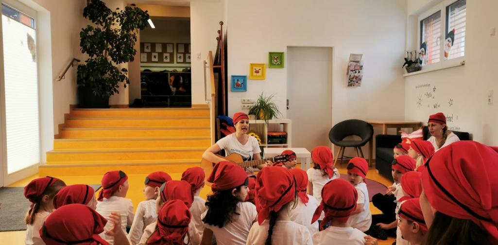 Verena Eder Eine Frau ist als Piratin verkleidet( mit rotem Kopftuch) und spielt auf der Gitarre. Viele Kinder mit rotem Kopftuch schauen zu ihr