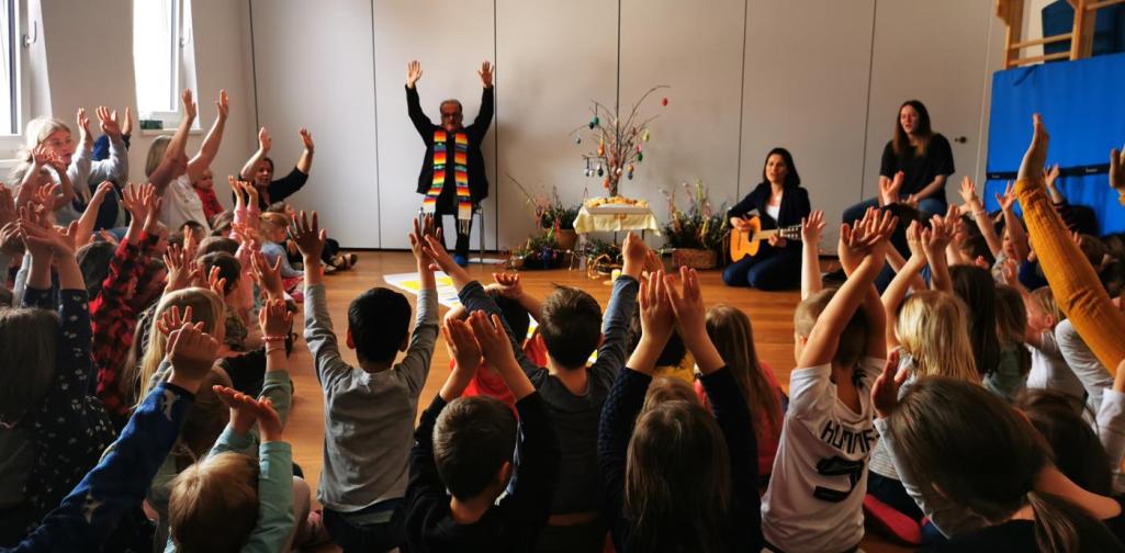Verena Der Pfarrer und viele Kinder, die man von hinten sieht, singen ein Lied und halten die Hände in die Höhe