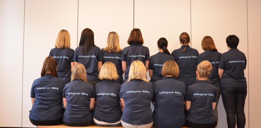 Das Kindergartenteam von hinten fotografiert. Alle haben das selbe T-Shirt mit der Aufschrift am Rücken: Kindergarten Polling an