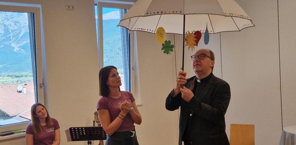Verena Eder Wünsche der Kinder hängen mit Symbolen am Schirm, welchen der Bischof über sich hält. Barbara und Stephanie betrachten den lachenden Bischof