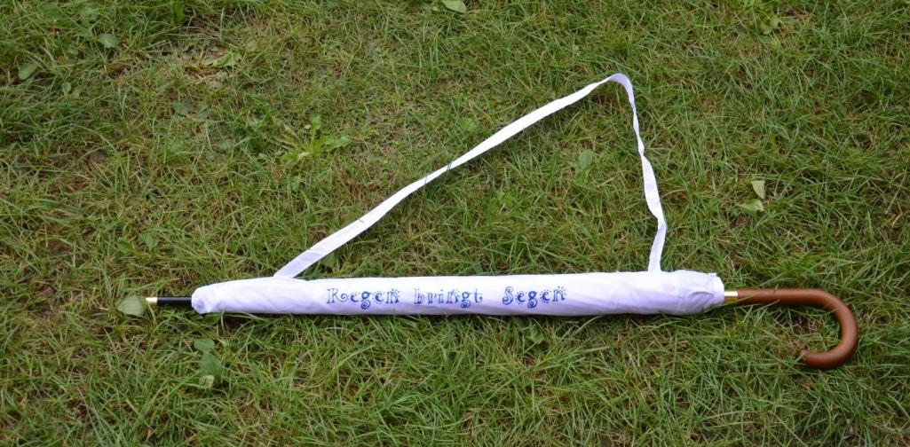 Der Regenschirm in seiner Hülle liegt auf einer Wiese. Die Hülle beinhaltet den Spruch &quot;Regen bringt Segen&quot;