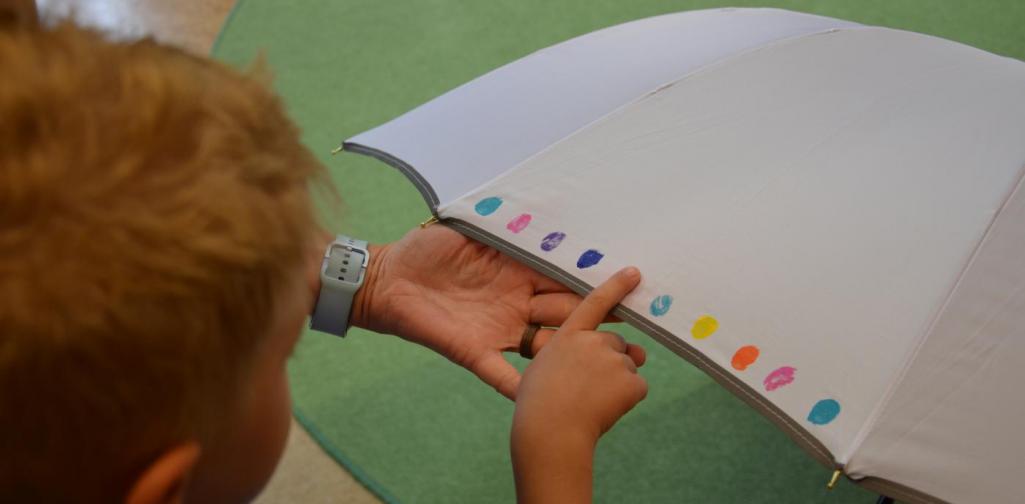 Über den Rücken des Kindes fotografiert, wie es einen bunten Fingerabdruck auf einem Weißen Regenschirm macht