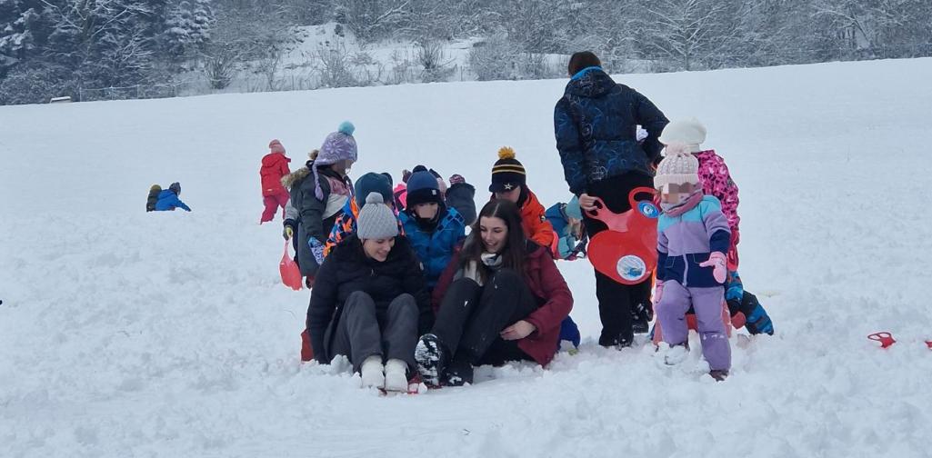 Verena Eder Zwei Erwachsene rutschen nebeneinander mit dem Rutschblatt- Kinder schieben sie an, damit sie eine Spur im frischen Schnee bekommen