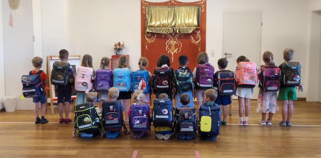 Alle Schulkinder des Jahres 2025/2026 stehen verkehrt in einer Reihe und zeigen ihre Schultaschen