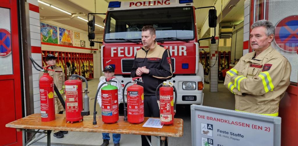 Zwei Feuerwehrleute stehen vor dem Feuerwehrauto und vor ihnen ist ein Bierbanktisch mit den verschiedensten Feuerlöschern zu sehen