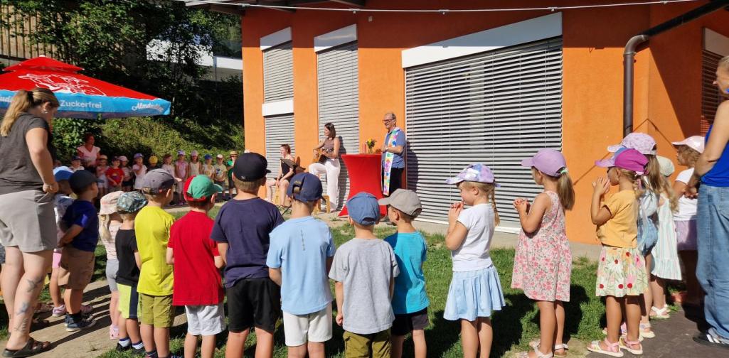 Zu sehen ist der Garten des Kindergartens. Die Kinder bilden einen Halbkreis um den Herr Pfarrer, der an der Kindergartenhauswand neben einem Stehtisch steht. Neben ihm steht Barbara mit der Gitarrer